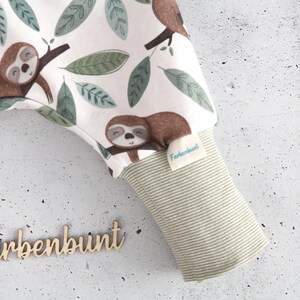May include: White fabric pants featuring a repeating pattern of brown sloths and green leaves. The garment includes a light green and white striped ribbed cuff with a small white tag labeled Farbenbunt. The word Farbenbunt is also displayed in wooden script.