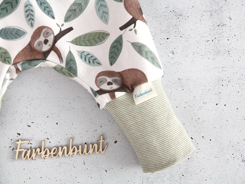 May include: White fabric pants featuring a repeating pattern of brown sloths and green leaves. The garment includes a light green and white striped ribbed cuff with a small white tag labeled Farbenbunt. The word Farbenbunt is also displayed in wooden script.