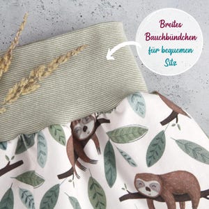 May include: A pair of pants featuring a white fabric with a repeating pattern of brown sloths on tree branches and green leaves. The pants have a wide, ribbed, sage green waistband. Text on the image reads: Breites Bauchb&uuml;ndchen f&uuml;r bequemen Sitz.