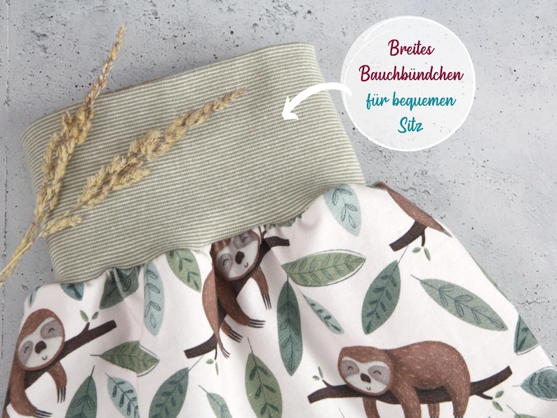 May include: A pair of pants featuring a white fabric with a repeating pattern of brown sloths on tree branches and green leaves. The pants have a wide, ribbed, sage green waistband. Text on the image reads: Breites Bauchb&uuml;ndchen f&uuml;r bequemen Sitz.