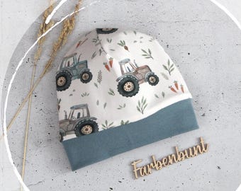 May include: A beanie hat featuring a white background with a repeating pattern of tractors, carrots, and green leaves. The hat has a solid blue-gray folded brim. A wooden sign with the text Farbenbunt is placed near the hat.