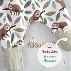 May include: Baby pants featuring a white background with a repeating pattern of brown sloths on tree branches and green leaves. The pants have long, ribbed, light green cuffs at the ankles and a small fabric tag labeled Farbenbunt on the right leg.