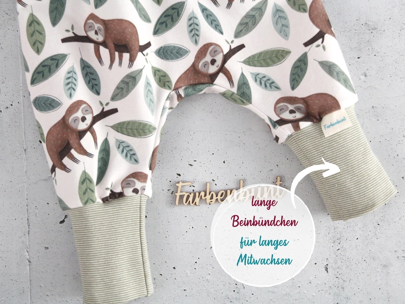 May include: Baby pants featuring a white background with a repeating pattern of brown sloths on tree branches and green leaves. The pants have long, ribbed, light green cuffs at the ankles and a small fabric tag labeled Farbenbunt on the right leg.