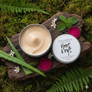 May include: A skincare product, "Beet Drift", is displayed in an open silver tin, revealing a creamy, light beige substance. The product is on a wooden surface with aloe vera, mint, and beet slices. The product is 2 oz / 57g.