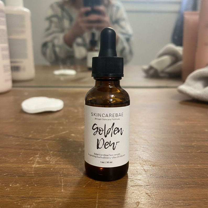 May include: A brown glass bottle of Skincarebae Golden Dew face serum with a black dropper. The label reads "Golden Dew" and "1 oz | 30 ml". The bottle is on a wooden surface.