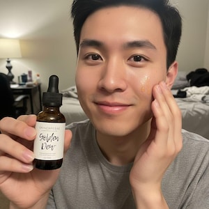 May include: A person holding a brown glass bottle of skincare product labeled "Golden Dew" by Skincare Bae. The person is applying the product to their face. The bottle has a black dropper and a white label with black text.