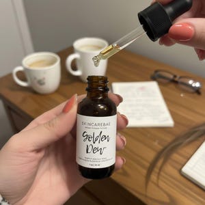 May include: A brown glass bottle of "Golden Dew" face serum with a dropper. The label reads "SKINCAREBAE" and "Botanical glow face serum". The dropper is dispensing the serum. The bottle contains 1 fl oz / 30 ml.
