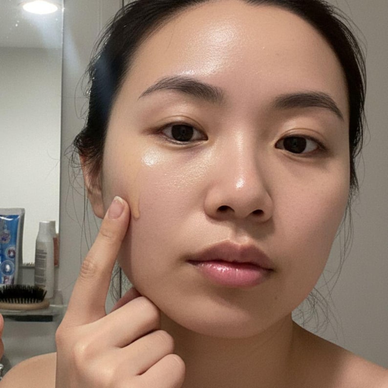 May include: A person applying a liquid cosmetic product to their face. The product is a light beige color and is being applied with a finger. The person is in a well-lit bathroom with a mirror in the background.
