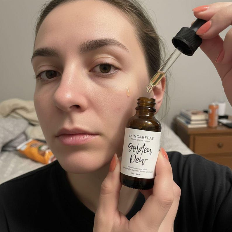 May include: A woman holding a brown glass bottle of skincare serum with a dropper. The bottle has a white label with the text "SKINCAREBAE" and "Golden Dew". A drop of serum is visible on the woman's face.