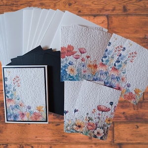 Puede incluir: Un conjunto de tarjetas en blanco con dise&ntilde;os florales. Las tarjetas son blancas con un patr&oacute;n en relieve y presentan ilustraciones florales de acuarela en tonos azules, rosas, naranjas y amarillos. El conjunto incluye respaldo de cartulina negra y sobres blancos.