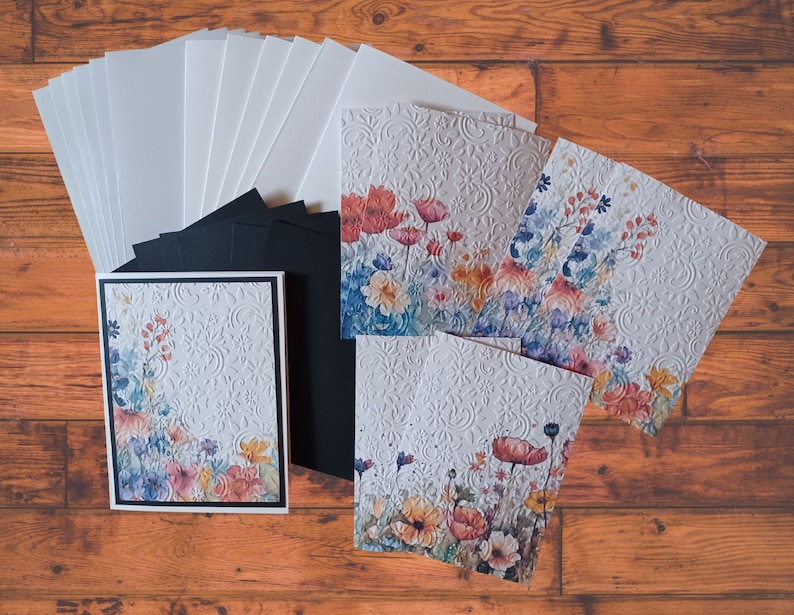 Puede incluir: Un conjunto de tarjetas en blanco con dise&ntilde;os florales. Las tarjetas son blancas con un patr&oacute;n en relieve y presentan ilustraciones florales de acuarela en tonos azules, rosas, naranjas y amarillos. El conjunto incluye respaldo de cartulina negra y sobres blancos.