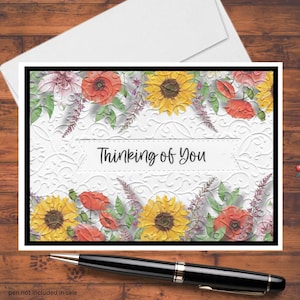 May include: A floral greeting card with the text "Thinking of You." The card features embossed white floral patterns and is bordered by sunflowers, poppies, and other flowers. A black pen rests below the card.