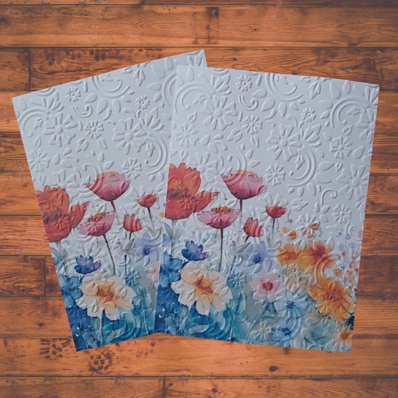 Puede incluir: Dos tarjetas blancas con un dise&ntilde;o floral. Las tarjetas presentan flores de acuarela en tonos de rosa, rojo y azul. El fondo es un patr&oacute;n en relieve blanco.