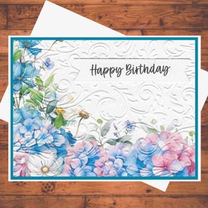 May include: A white greeting card with a teal border and a floral design. The card features blue and pink hydrangeas and the words "Happy Birthday" in black script. The background has a subtle embossed pattern.