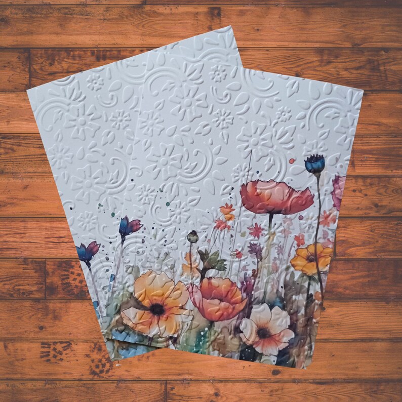 Puede incluir: Una tarjeta blanca con un patr&oacute;n floral en relieve y una pintura de acuarela de un campo de flores en tonos rojos, naranjas, amarillos y azules.