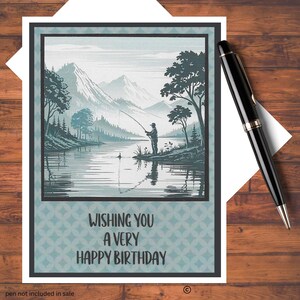 May include: A birthday card with a serene landscape illustration of a person fishing by a lake, mountains in the background. The card has the text "WISHING YOU A VERY HAPPY BIRTHDAY" in black. A black pen rests on the wooden surface.