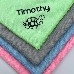 May include: A stack of four microfiber cloths in pink, blue, grey, and green. The green cloth has the name "Timothy" embroidered on it, along with a black and white turtle design.