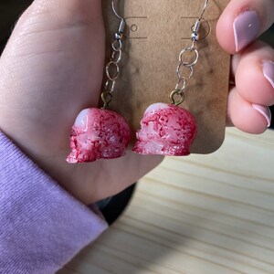 K&ouml;nnte beinhalten: Ein Paar silberne Sch&auml;del-Ohrringe mit einem roten und wei&szlig;en Farbmuster. Die Sch&auml;del sind detailliert und haben ein realistisches Aussehen.
