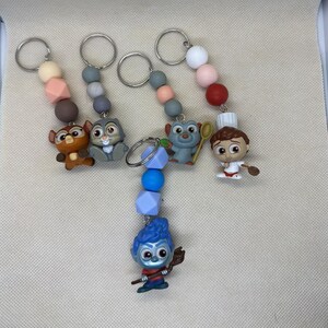 Puede incluir: Llaveros con figuras de personajes de dibujos animados. Cada llavero tiene un anillo de metal, una cadena de cuentas de colores y una pequeña figura de personaje. Los personajes incluyen un búho, un conejo, una criatura azul y un chef. Las cuentas son en tonos rosa, gris y azul.