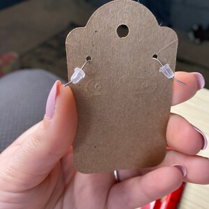 K&ouml;nnte beinhalten: Eine braune Papier-Ohrringkarte mit zwei silbernen Ohrringpfosten und durchsichtigen Kunststoff-Ohrringr&uuml;ckseiten.