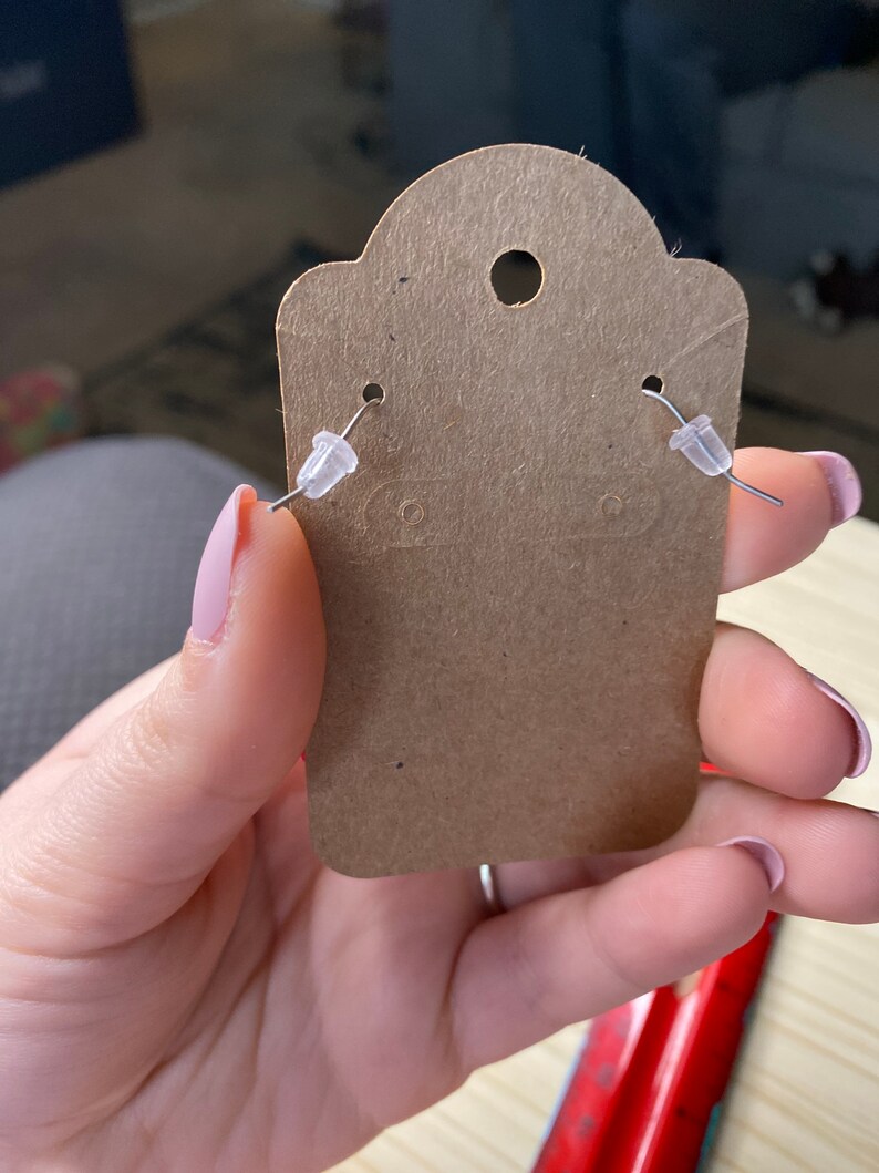 K&ouml;nnte beinhalten: Eine braune Papier-Ohrringkarte mit zwei silbernen Ohrringpfosten und durchsichtigen Kunststoff-Ohrringr&uuml;ckseiten.