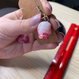 K&ouml;nnte beinhalten: Ein Paar silberne Sch&auml;del-Ohrringe mit einer wei&szlig;en Basis und roten Farbstichen. Die Ohrringe h&auml;ngen an einer braunen Karte.