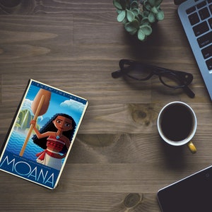 May include: A book cover featuring a cartoon illustration of a Polynesian woman with brown hair and a red and white outfit holding a wooden paddle. The book cover is blue and white with the text "Moana" in large blue letters and the text "Finding the Way" in smaller white letters.