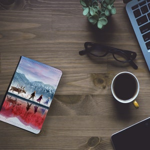 May include: A leather-bound notebook with a colorful image of a snowy landscape and characters from the movie Frozen. The characters are walking on a frozen lake with a reindeer.