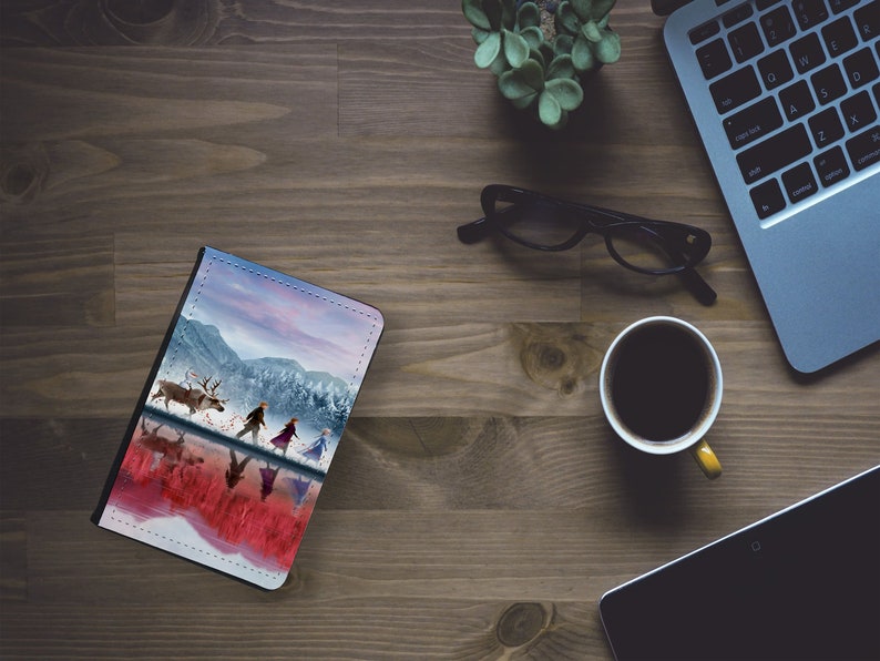 May include: A leather-bound notebook with a colorful image of a snowy landscape and characters from the movie Frozen. The characters are walking on a frozen lake with a reindeer.