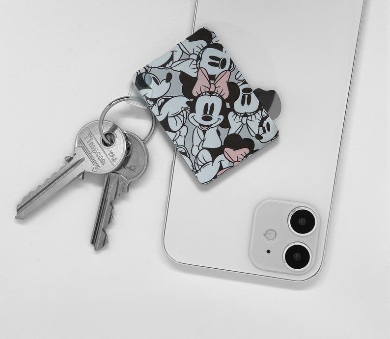 May include: A white smartphone with a black camera lens and a Mickey Mouse and Minnie Mouse character phone grip attached to the back.