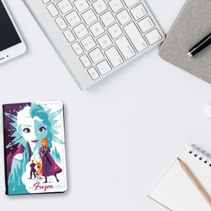 May include: A white and purple passport holder with a Frozen design featuring Elsa and Anna. The design includes a snowflake pattern and the word "Frozen" in pink letters.