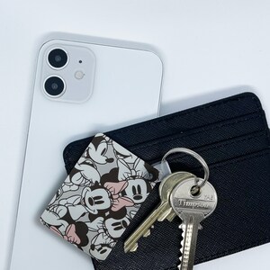 May include: A black leather wallet with a Mickey Mouse character print card holder. The card holder features a variety of Mickey Mouse characters in black and white. The wallet has a silver key chain with two keys attached. One key has the text "YALE" and the other key has the text "TIMPSON".