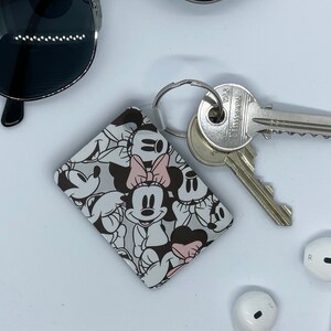 May include: A white and black keychain with a Mickey Mouse and Minnie Mouse design. The keychain has a silver ring and is attached to a set of keys.