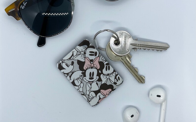 May include: A white and black keychain with a Mickey Mouse and Minnie Mouse design. The keychain has a silver ring and is attached to a set of keys.
