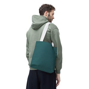Può includere: Una borsa tote color teal con una tracolla bianca viene portata sulla spalla. La borsa è di colore pieno e sembra essere fatta di un tessuto resistente. La persona indossa una felpa con cappuccio verde e pantaloni scuri.