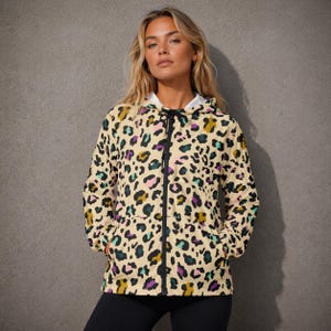 Peut inclure: Veste à capuche imprimé léopard beige avec des taches noires, violettes, turquoise et dorées. La veste a une fermeture éclair noire et une doublure blanche dans la capuche. Le modèle porte un legging noir.
