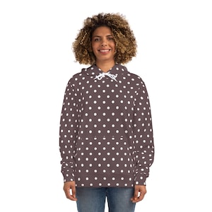 Può includere: Una felpa con cappuccio marrone con un motivo a pois bianchi. La felpa ha un cappuccio con coulisse e una tasca frontale. La modella indossa jeans blu.