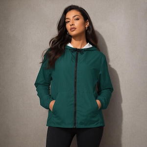 Puede incluir: Chaqueta con capucha verde oscuro con cremallera negra y dos bolsillos delanteros. La chaqueta tiene un forro blanco en la capucha y se usa sobre pantalones negros. La modelo está de pie frente a una pared gris.