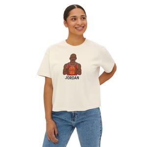Può includere: T-shirt color crema con un'illustrazione a cartoni animati di un giocatore di basket che tiene in mano un pallone. L'illustrazione è in rosso, marrone e nero, con il nome "JORDAN" stampato sotto. La maglietta è indossata da una persona.
