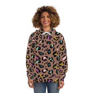 Può includere: Una felpa con cappuccio marrone con stampa leopardata colorata. Il motivo include macchie nere, gialle, viola e turchese. La felpa ha un cordoncino e una tasca frontale. La modella indossa jeans blu.