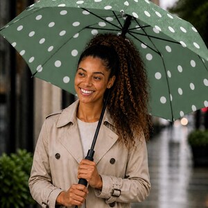 May include: A green umbrella with white polka dots is held open. The umbrella is held by a person wearing a tan trench coat. The background is blurred, suggesting an outdoor setting, possibly a rainy day.