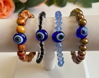 May include: Four bracelets with blue evil eye charms. The bracelets are made of different materials, including wood, black beads, blue crystal beads, and iridescent brown beads.