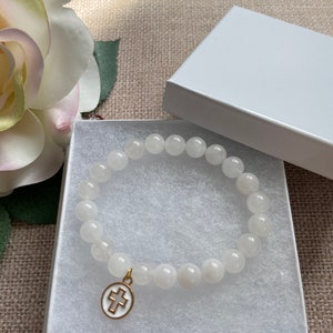 Op de afbeelding: Witte kralenarmband met een goudkleurige kruisbedel, gepresenteerd in een witte geschenkdoos. De armband heeft ronde, ondoorzichtige kralen. De kruisbedel heeft een wit midden en een goudkleurige rand.