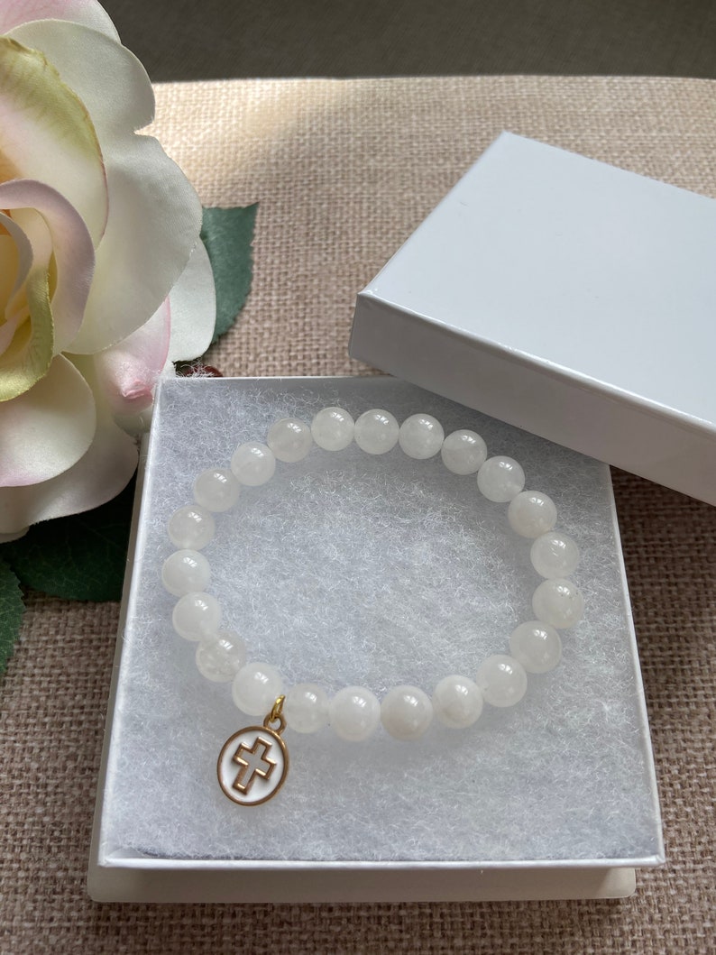 Op de afbeelding: Witte kralenarmband met een goudkleurige kruisbedel, gepresenteerd in een witte geschenkdoos. De armband heeft ronde, ondoorzichtige kralen. De kruisbedel heeft een wit midden en een goudkleurige rand.