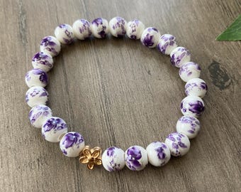 Pode incluir: Pulseira de cerâmica branca com desenhos florais roxos e um fecho de flor dourado.