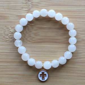 Op de afbeelding: Witte kralenarmband met een goudkleurige kruisbedel. De armband heeft ronde, ondoorzichtige witte kralen. De bedel is een goudkleurige cirkel met een wit kruis in het midden. Op een lichtbruin houten oppervlak.