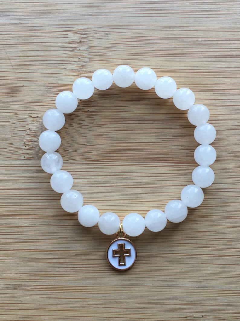 Op de afbeelding: Witte kralenarmband met een goudkleurige kruisbedel. De armband heeft ronde, ondoorzichtige witte kralen. De bedel is een goudkleurige cirkel met een wit kruis in het midden. Op een lichtbruin houten oppervlak.