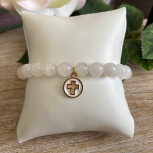 Op de afbeelding: Witte kralenarmband met een goudkleurige kruisbedel. De armband is gemaakt van ronde, lichtgekleurde kralen en wordt gepresenteerd op een wit kussen. De kruisbedel heeft een wit midden en een goudkleurige rand.