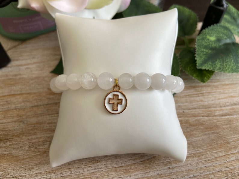 Op de afbeelding: Witte kralenarmband met een goudkleurige kruisbedel. De armband is gemaakt van ronde, lichtgekleurde kralen en wordt gepresenteerd op een wit kussen. De kruisbedel heeft een wit midden en een goudkleurige rand.