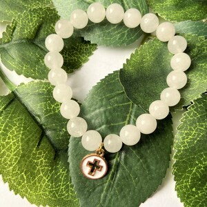 Op de afbeelding: Een armband met ronde, cr&egrave;mekleurige kralen. Een goudkleurige bedel met een witte emaille achtergrond en een zwart kruis hangt aan de armband. De armband ligt op groene bladeren.